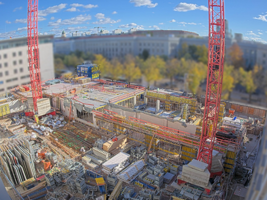 Das Bild zeigt eine große Baustelle inmitten eines städtischen Umfelds, aufgenommen aus erhöhter Perspektive. Im Zentrum der Szene befinden sich zwei auffällige rote Baukräne, die senkrecht in die Höhe ragen und den Blick dominieren. Auf der Baustelle selbst sind zahlreiche Baumaterialien wie Stahlträger, Holzverschalungen, Betonplatten und Gerüste ordentlich gestapelt. Der Rohbau ist bereits weit fortgeschritten: Teile des Fundaments und erste Wände sind sichtbar, was auf den Bau eines mehrstöckigen Gebäudes hindeutet.