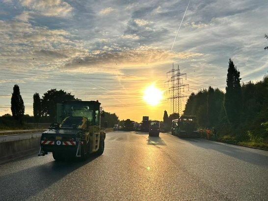 Straßenbauarbeiten bei Sonnenaufgang auf einer mehrspurigen Fahrbahn. Mehrere Baumaschinen und Fahrzeuge sind auf der frisch asphaltierten Straße verteilt. Im Hintergrund sind Strommasten und Bäume zu sehen, der Himmel ist teils bewölkt mit sichtbaren Kondensstreifen.
