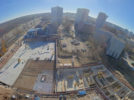 Das Bild zeigt eine Luftaufnahme einer großen Baustelle aus erhöhter Perspektive, vermutlich aufgenommen von einem Kran oder einer fest installierten Baustellenkamera. Im Zentrum ist ein rechteckiges Baufeld zu sehen, auf dem bereits umfangreiche Betonarbeiten ausgeführt wurden. Große, helle Betonflächen deuten auf eine fertiggestellte Bodenplatte bzw. den Rohbau der Weißen Wanne hin. In Teilbereichen sind Schalungen, Bewehrungen und einzelne aufgehende Bauteile erkennbar. Rechts im Bild befinden sich strukturierte Gruben und Fundamentbereiche, die sich noch in einem früheren Bauzustand befinden. Das Baufeld ist von Baustraßen, Absperrungen und Baumaterialien umgeben. Am linken Bildrand ist ein gelber Kranarm sichtbar, der die Perspektive und den aktiven Hochbaukontext unterstreicht.