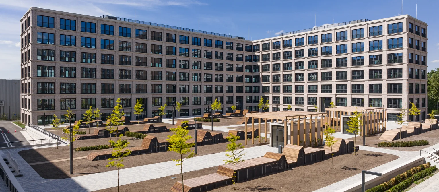 TechnoCampus, Berlin View of the TechnoCampus; in the foreground you can see many wooden seats; there are several trees planted; in between are footpaths; in the background is the L-shaped building with the extensive glass front and sandstone-coloured façade.
