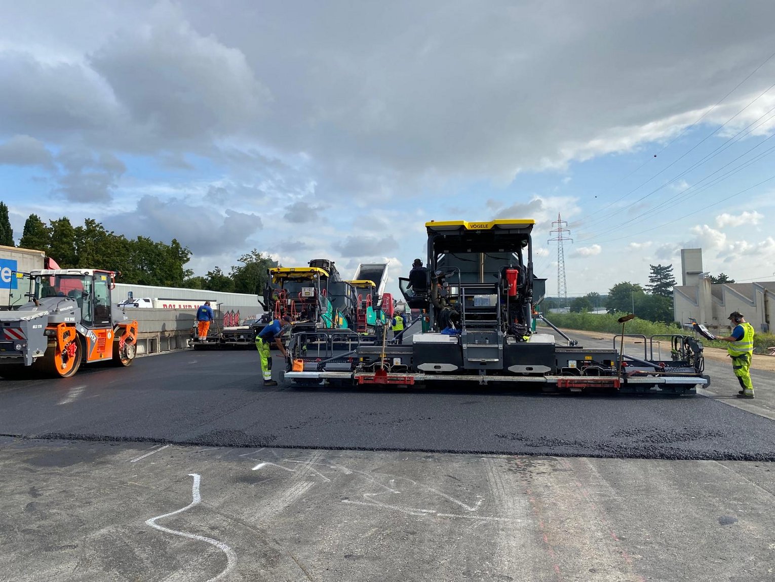 Das Bild zeigt eine Straßenszene während einer Bauphase. Im Mittelpunkt stehen drei große Asphaltfertiger, die frischen Asphalt auf die Straße auftragen. Links davon befindet sich eine Straßenwalze mit orangefarbenen Rädern, die zur Verdichtung des Asphalts eingesetzt wird. Mehrere Arbeiter in Warnkleidung sind rund um die Maschinen aktiv mit der Straßenarbeit beschäftigt. Der Himmel ist teilweise bewölkt, und im Hintergrund sind Bäume sowie weitere Infrastruktur zu erkennen.