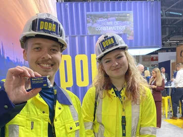 Zwei Bauarbeiter mit Sicherheitsklamotten machen ein Foto auf der Messe. Der vordere hält eine Mannerschnitte in die Höhe.