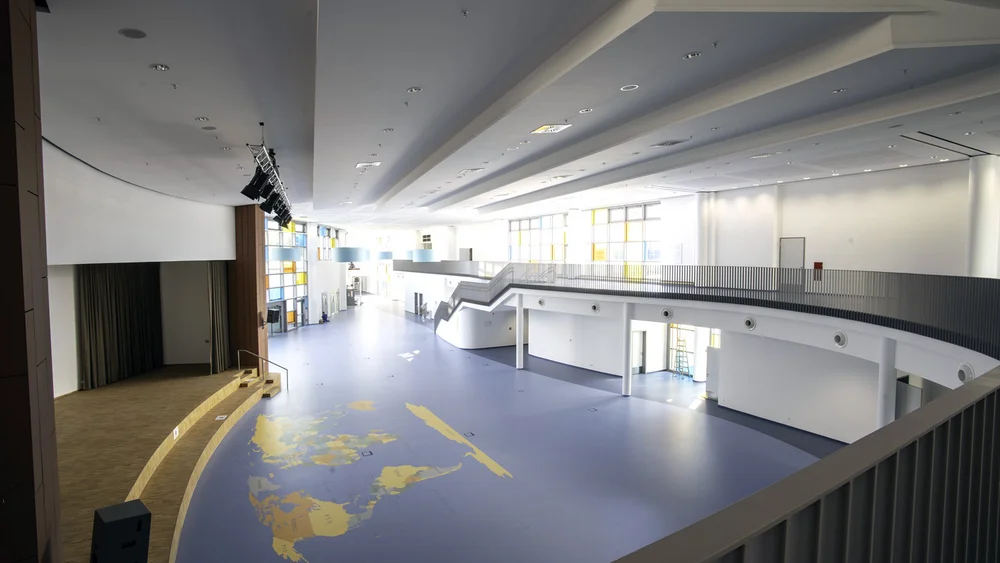 Foto: Blick von einer Galerie in einen langgezogenen, hellen Raum mit großer Weltkarte auf blauem Boden, links eine Bühne, im Hintergrund bunte Fenster