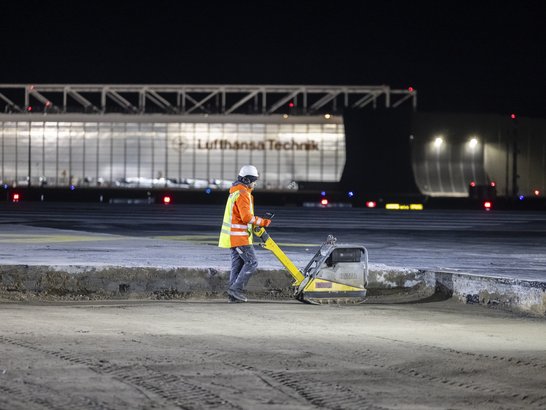 Nächtliche Sanierungsarbeiten auf dem Flughafen Frankfurt: Eine Fachkraft verdichtet den Untergrund einer Verkehrsfläche mit einer Rüttelplatte vor dem Einbau einer neuen Beton- oder Asphaltdecke. Im Hintergrund ist ein Hangar der Lufthansa Technik zu sehen.