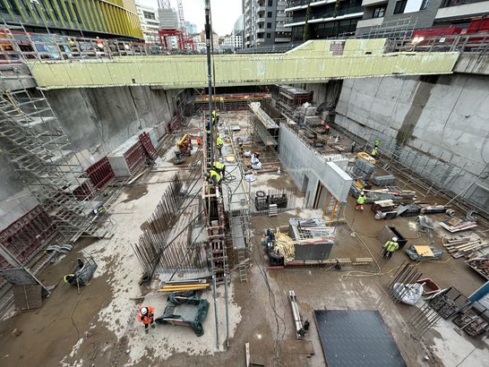Die Aufnahme zeigt eine große Baugrube, die sich in einem fortgeschrittenen Stadium des Rohbaus befindet. Mehrere Ebenen sind bereits durch massive Betonwände und Zwischendecken strukturiert. Auffällig sind die zahlreichen Bewehrungsstäbe, die aus den Wänden herausragen und auf bevorstehende Betonarbeiten hinweisen. Auf der rechten Seite befindet sich eine gelbe Schalungsfläche, die für die nächste Bauphase vorbereitet wurde. Links im Bild ist ein mehrstöckiges Gerüst installiert, das den Zugang zu den unterschiedlichen Ebenen ermöglicht. Am Boden liegen verschiedene Baumaterialien wie Stahlträger und Holzbohlen, ergänzt durch Maschinen und Werkzeuge, die für den weiteren Bauablauf benötigt werden. Sicherheitsmaßnahmen sind ebenfalls erkennbar: Am oberen Rand der Baugrube sind Schutzgeländer angebracht, um Abstürze zu verhindern.