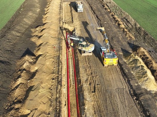 Baustelle eines tief ausgehobenen Kabelgrabens auf einem Feld. Mehrere Baumaschinen arbeiten entlang der Trasse, darunter ein Bagger und ein Fahrzeug mit Kranaufsatz. Rote Schutzrohre liegen im offenen Graben, während Erde auf beiden Seiten aufgeschüttet ist.
