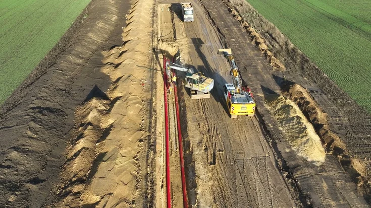 Baustelle eines tief ausgehobenen Kabelgrabens auf einem Feld. Mehrere Baumaschinen arbeiten entlang der Trasse, darunter ein Bagger und ein Fahrzeug mit Kranaufsatz. Rote Schutzrohre liegen im offenen Graben, während Erde auf beiden Seiten aufgeschüttet ist.