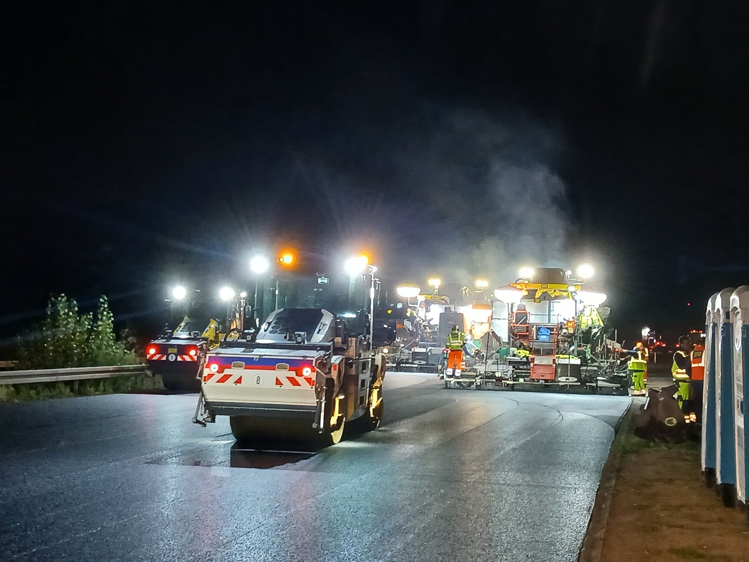 Nächtliche Autobahnbaustelle mit Asphaltfertigern, Walzen und Begleitfahrzeugen, die unter intensiver Beleuchtung eine frisch asphaltierte Fahrbahn einbauen; mehrere Baukräfte in Warnkleidung arbeiten entlang der Fahrbahn, rechts sind mobile Toilettenkabinen zu sehen, im Hintergrund steigen Dampf und Rauch aus dem heißen Asphalt auf.