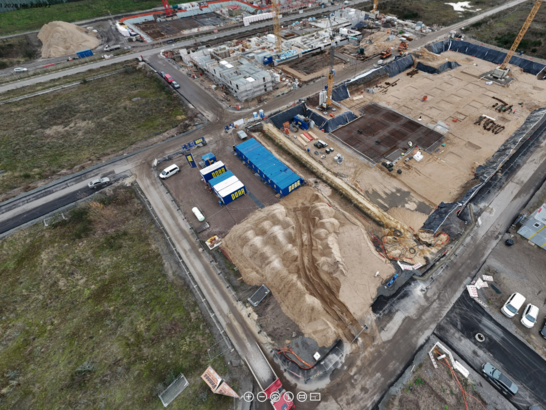 Luftaufnahme einer Hochbau‑Baustelle mit Baugrube, Bodenplatten, Kränen und Baustelleneinrichtung in der Gründungsphase.