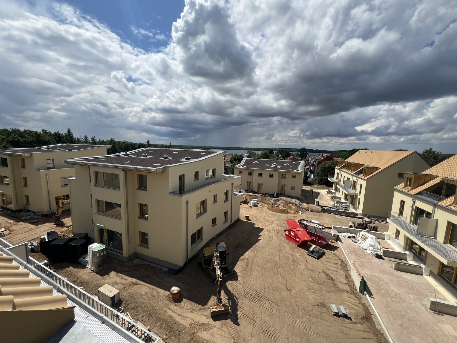 Das Bild zeigt eine Baustelle mit mehreren Gebäuden in unterschiedlichen Bauphasen. Die Gebäude sind hellgelb und haben sowohl Flach- als auch Schrägdächer. Auf dem Gelände befinden sich Baumaterialien und ein kleiner Bagger. Der Himmel ist teilweise bewölkt, mit sichtbaren blauen Stellen.