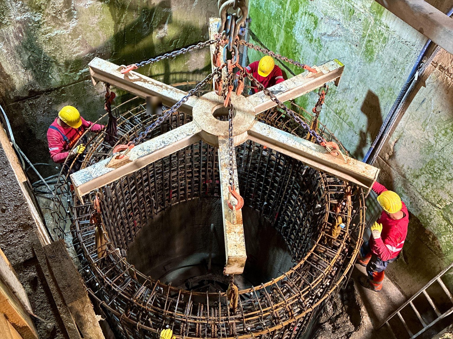 Drei Bauarbeiter in Schutzausrüstung arbeiten an einer Stahlbewehrung für eine kreisförmige Betonkonstruktion, vermutlich ein Fundament oder Schacht, in einer Baugrube. Sie sind an einem Gerüst gesichert.