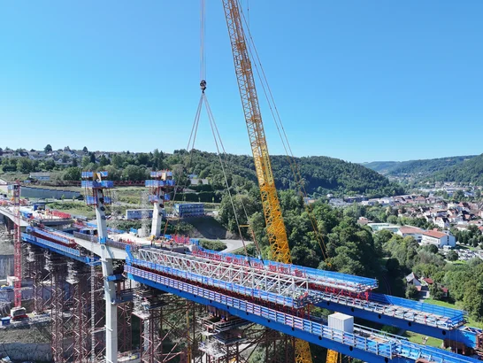 Large construction site for a bridge: A yellow crane lifts a large component over a valley while workers labor on the bridge. Green hills, a city, and blue scaffolding are visible in the background.