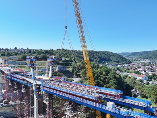 Großbaustelle für eine Brücke: Ein gelber Kran hebt ein großes Bauteil über ein Tal, während Arbeiter auf der Brücke arbeiten. Im Hintergrund sind grüne Hügel, eine Stadt und blaue Gerüste sichtbar.