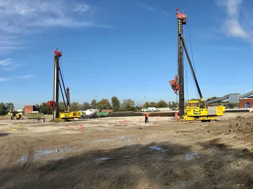 Zwei Spezialtiefbau rammen stehen auf einen großen Baufeld. Ein paar Arbeiter laufen über das Feld