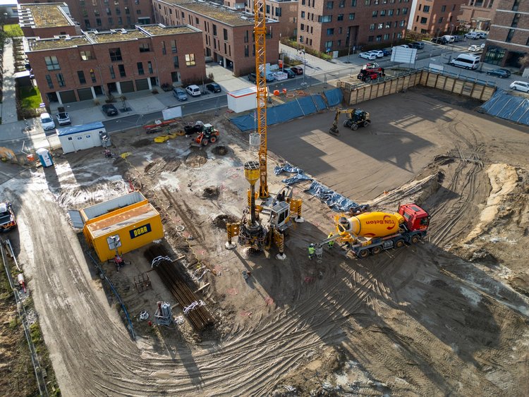 A construction site where an atlas pile is being installed. A large yellow drilling rig stands in the middle of the picture, surrounded by construction workers and various construction machines. A concrete mixer truck is to the right of the rig. Several buildings can be seen in the background.