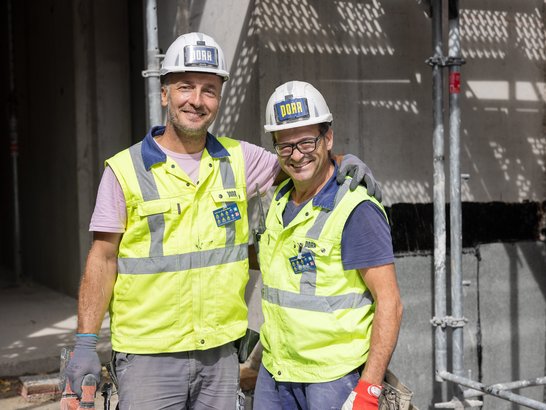 Auf dem Bild stehen zwei Bauarbeiter auf einer Baustelle dicht nebeneinander und haben freundschaftlich die Arme umeinandergelegt. Beide tragen helle Schutzhelme mit Firmenlogo sowie auffällige, neongelbe Warnwesten über Arbeitskleidung. An den Händen sind Arbeitshandschuhe zu sehen, und an den Westen befinden sich Ausweise oder Namensschilder.