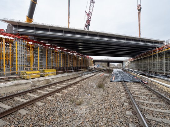 Luftaufnahme einer nächtlichen Baustelle in Marzahn mit beleuchteten Kränen und Baugeräten an einer Kreuzung von Straßen und Eisenbahnschienen, im Hintergrund die Lichter der Stadt.