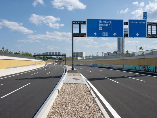 Das Bild zeigt eine neu gebaute Autobahn mit jeweils zwei Fahrspuren pro Richtung. In der Mitte befindet sich ein mit Kies gefüllter Mittelstreifen, der durch eine Metallbarriere getrennt ist. Über der Fahrbahn hängen zwei blaue Verkehrsschilder: Das linke Schild weist in Richtung Hamburg und Dresden, das rechte in Richtung Sonnenallee. Der Himmel ist klar mit einigen verstreuten Wolken, und im Hintergrund sind Gebäude sowie Grünflächen zu sehen.