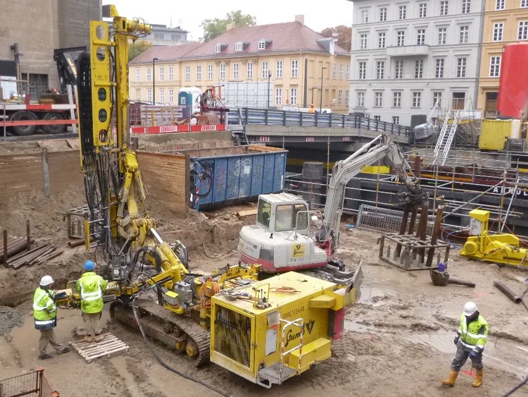 Foto: Die Baugrube mit zahlreichen kleineren Baugeräten. Im Vordergrund wird ein Pfahl gebohrt. Im Hintergrund werden Materialien von einer Position auf eine andere umgelagert.