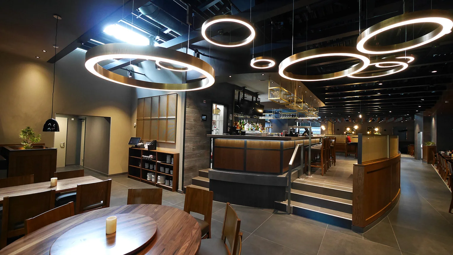Extravagant lighting. Photo: area in front of the bar with circular neon tubes hanging from the ceiling; wooden floor; two sets of three steps up to the bar area, a round table and a rectangular table with comfortable chairs at the left side of the picture