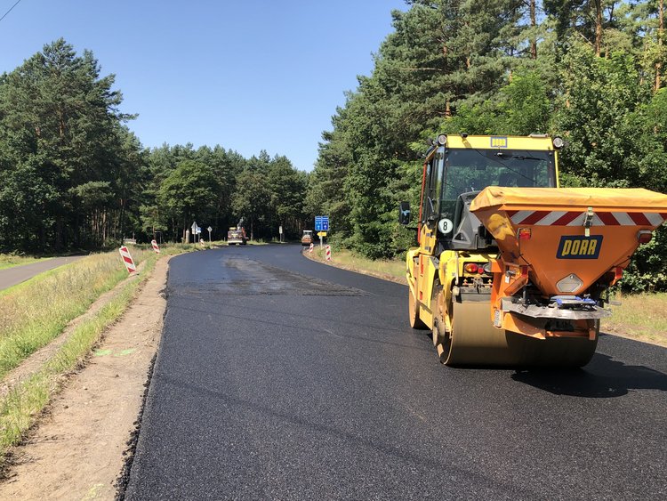 The picture shows a street scene during construction work. A yellow road roller marked ‘PORR’ is laying a new layer of asphalt on a rural road surrounded by trees and green spaces.