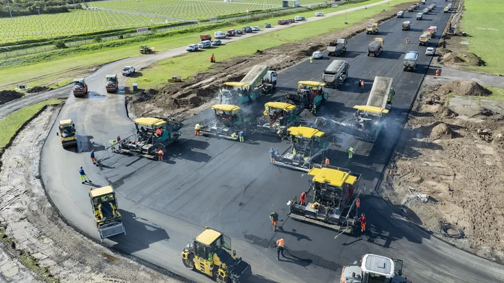 Das Bild zeigt eine groß angelegte Straßenbaustelle, auf der eine neue Fahrbahn entsteht. Mehrere gelbe Asphaltfertiger arbeiten nebeneinander und tragen frischen Asphalt auf die breite Fläche auf. Vor und hinter ihnen sind Walzen im Einsatz, die die Oberfläche verdichten. Zahlreiche LKW liefern das heiße Mischgut direkt zu den Maschinen, während Bauarbeiter in auffälligen Warnwesten die Arbeiten überwachen und koordinieren. Über den Fertigern steigt leichter Rauch auf, was auf den Einsatz von heißem Material hinweist. Die Baustelle befindet sich in einer offenen Landschaft mit grünen Feldern und landwirtschaftlichen Flächen im Hintergrund. Am Horizont sind Industrieanlagen und einige Gebäude zu erkennen.