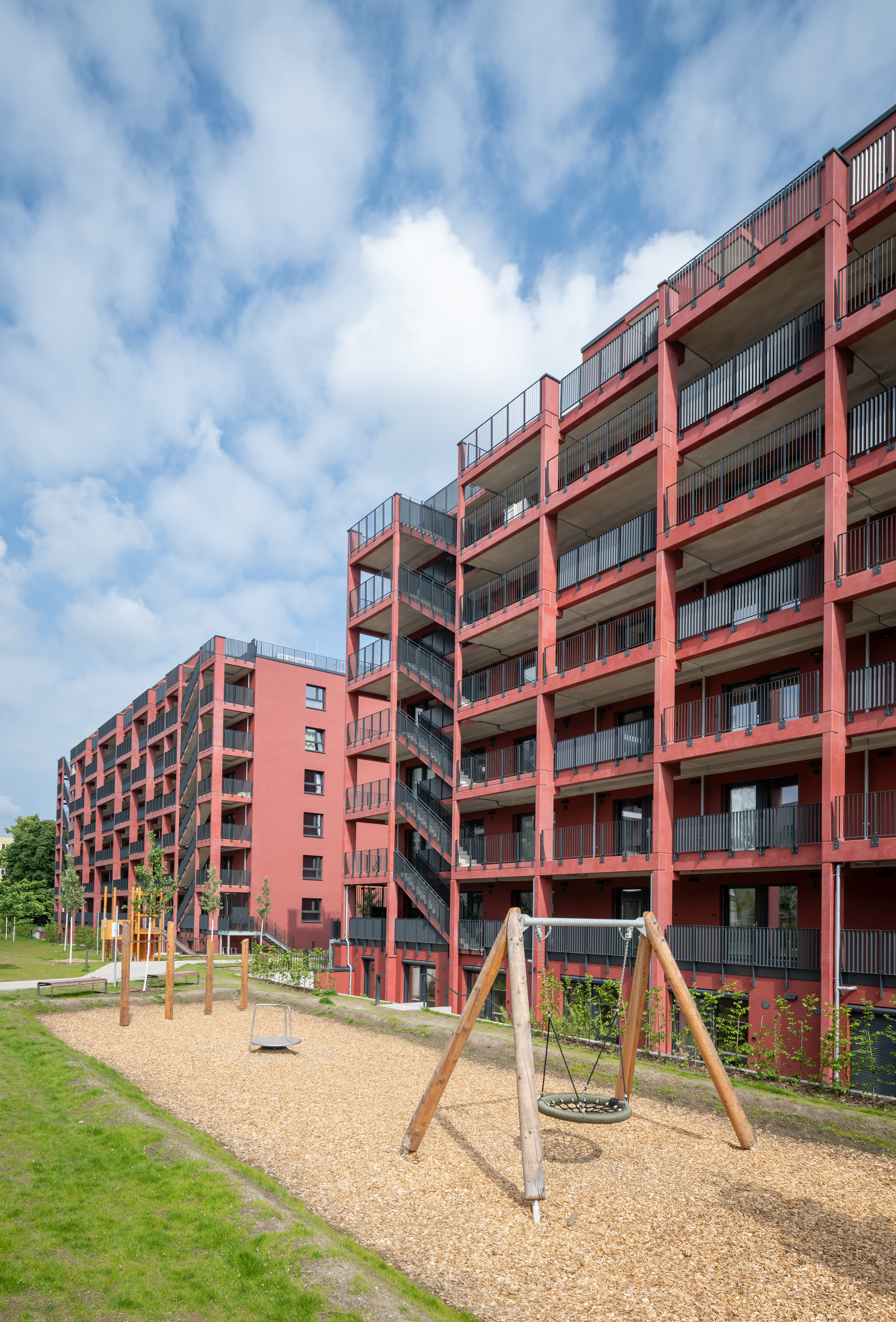 Blick auf einen Spielplatz im Sand und dahinter steht ein Gebäude mit roter Fassade.
