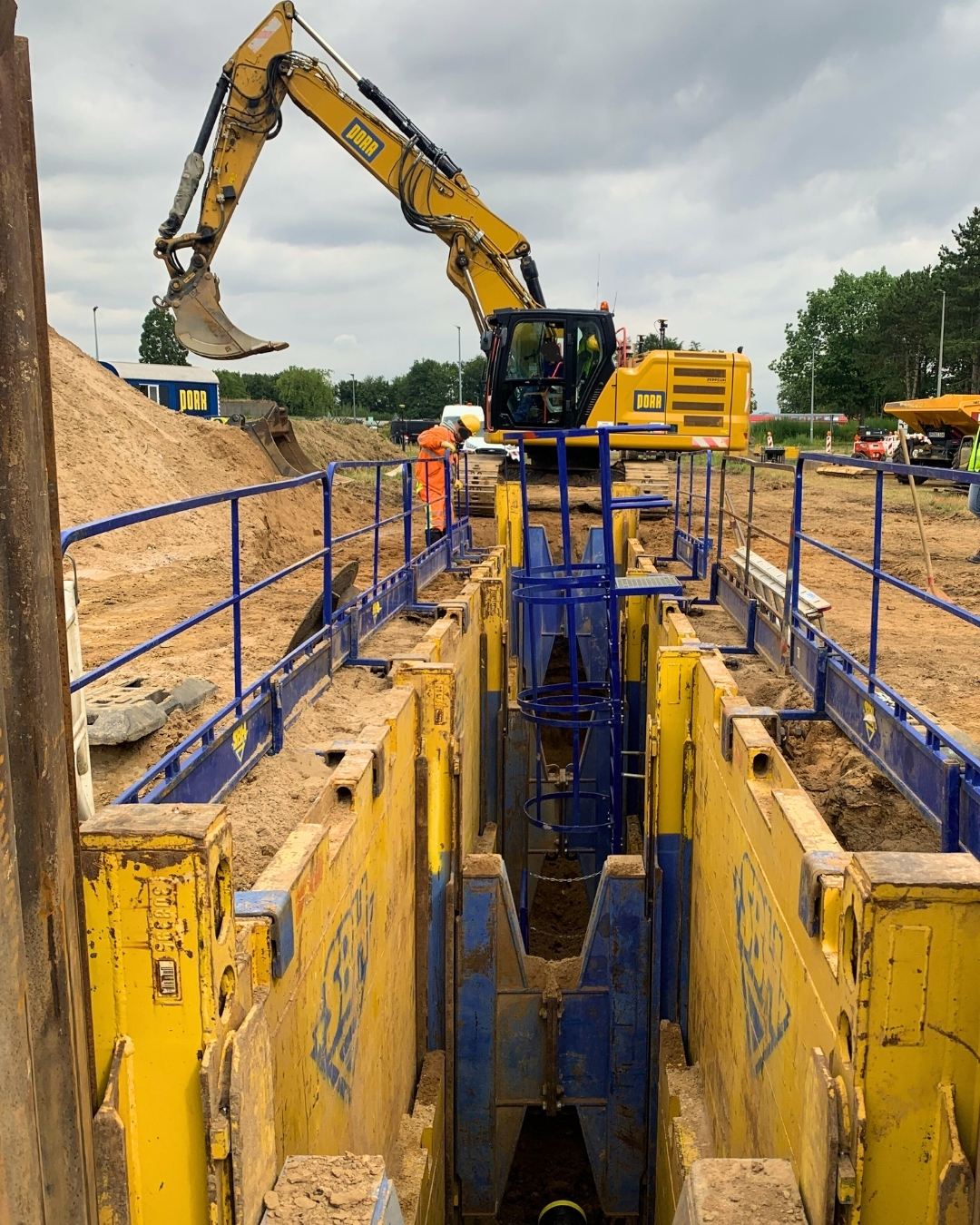 Construction site for pipeline construction with secured trench, excavator, and construction workers in protective clothing; surrounded by trees and construction machinery.